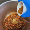 beans and tomatoes in large stock pot with gloved hand pouring in spices.