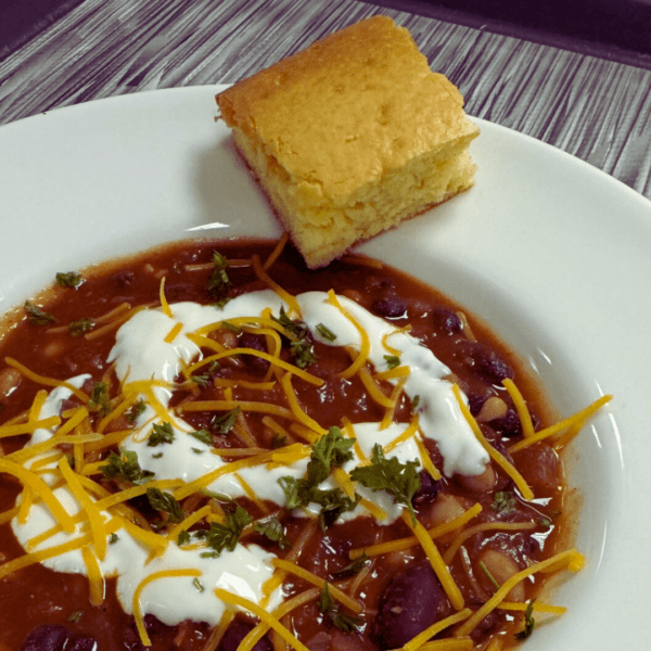 entree bowl with three bean chili topped with sour cream and cheddar cheese with a piece of corn bread.