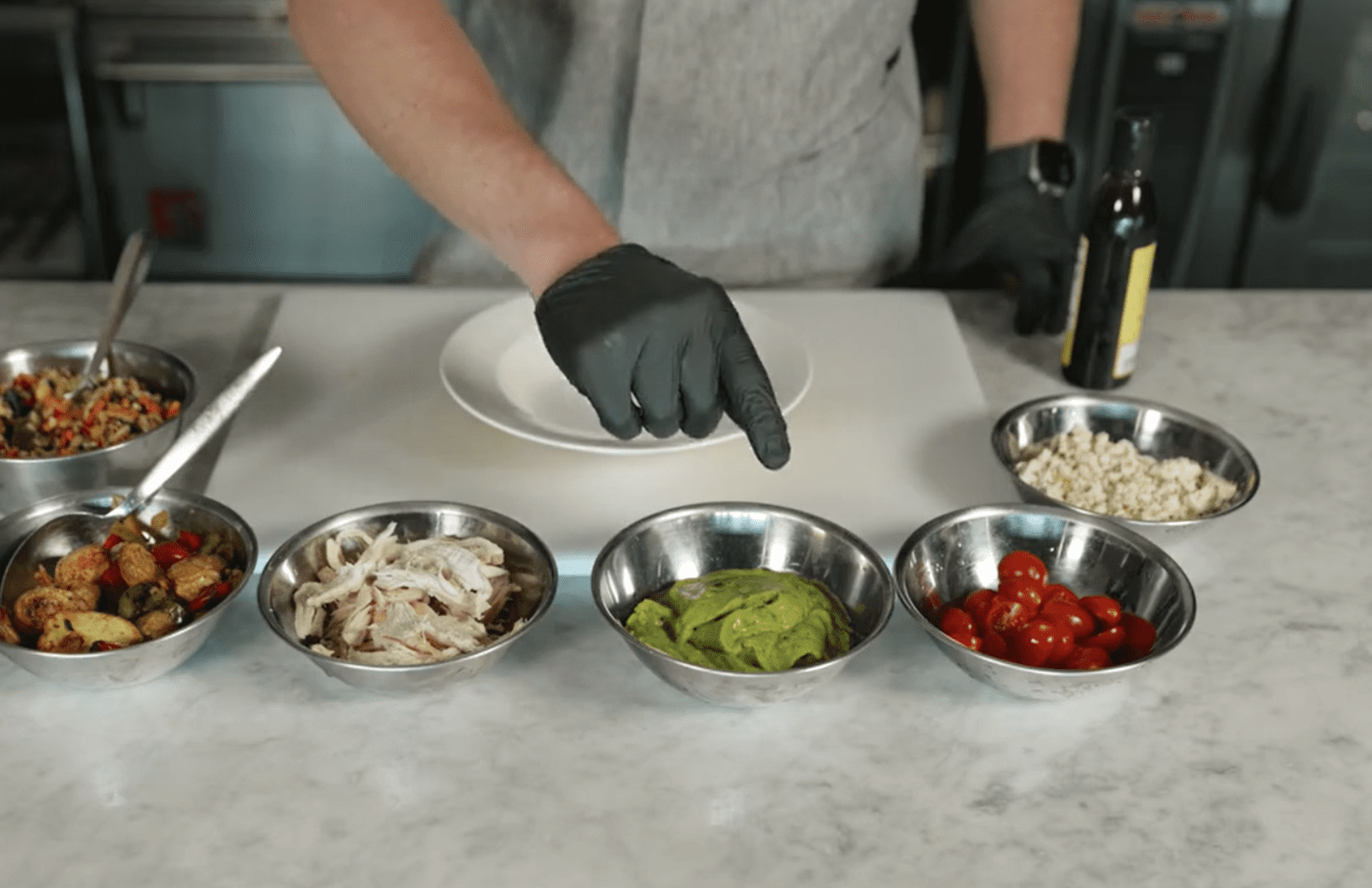 metal bowls on counter with various ingredients pre-portioned out while chef in black gloves points to bowl containing guacamole.